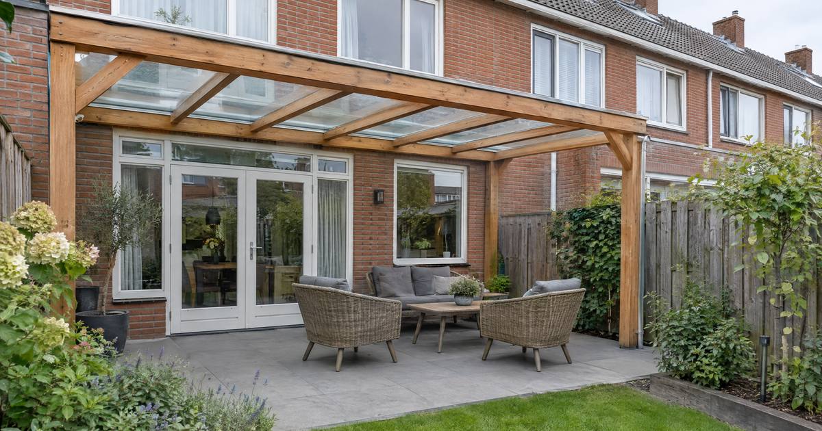 Een houten overkapping met een glazen dak boven een terras aan de achterkant van een rijtjeshuis, met tuinstoelen eronder. Illustratie.